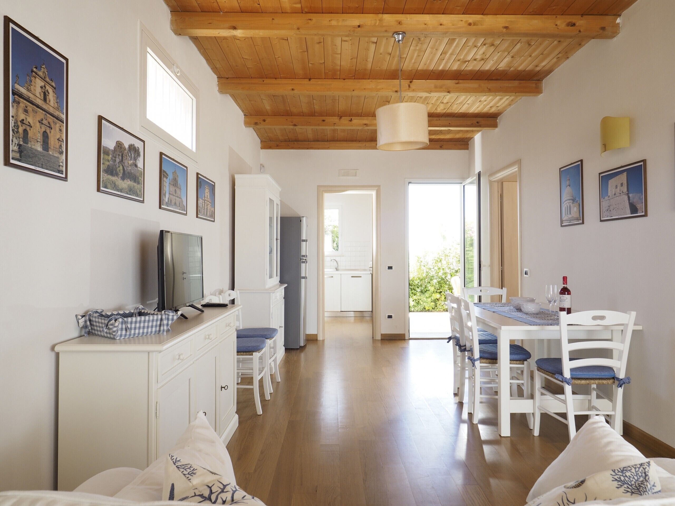 A spacious living area with a wooden ceiling, a dining table with chairs, a kitchen area, and a TV on a cabinet.