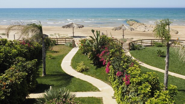 A landscaped garden with a winding path leading to a beach with thatched umbrellas.