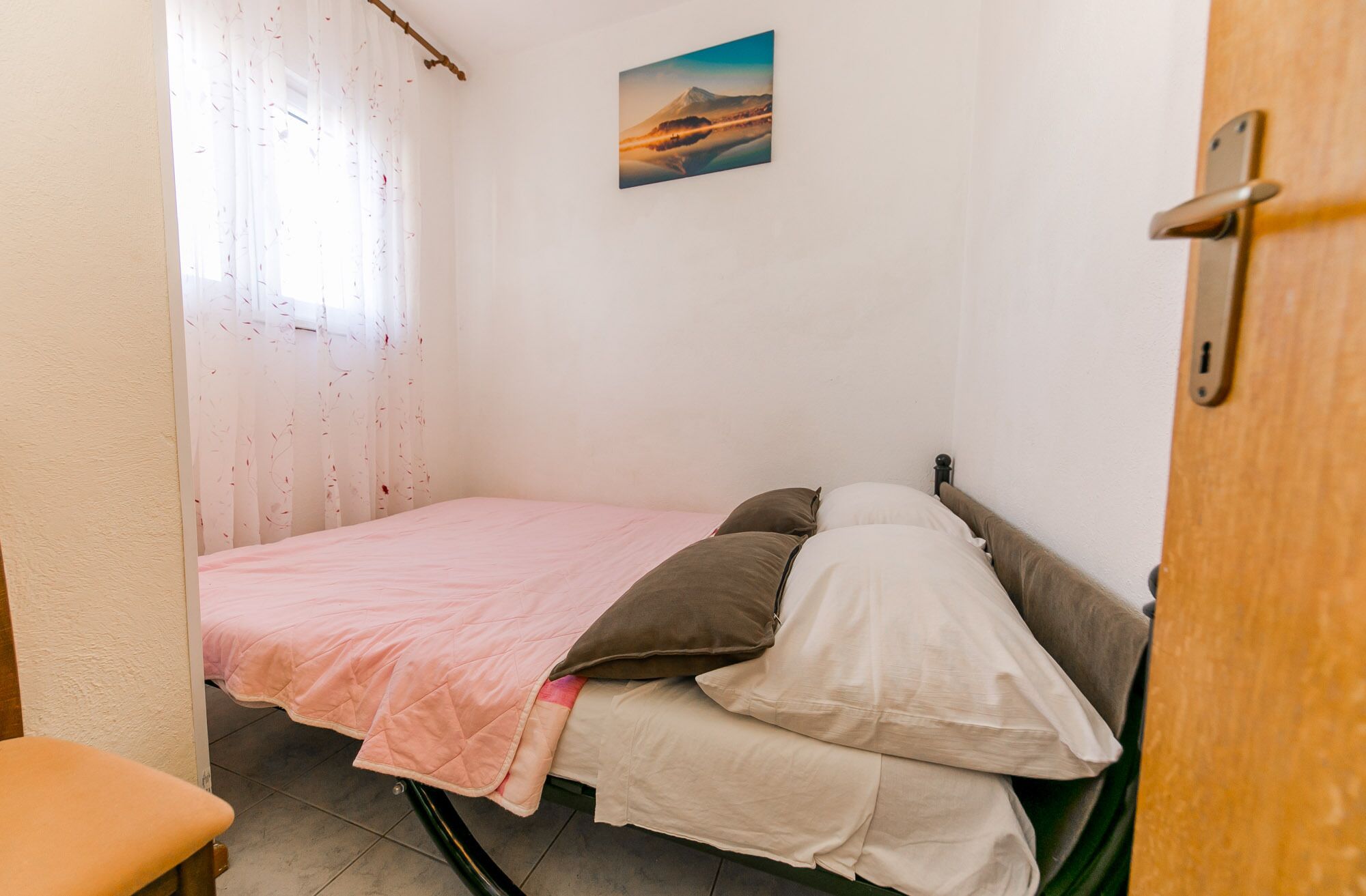 A small, single-bed room with a pink bedspread, a wooden door, and a painting on the wall.
