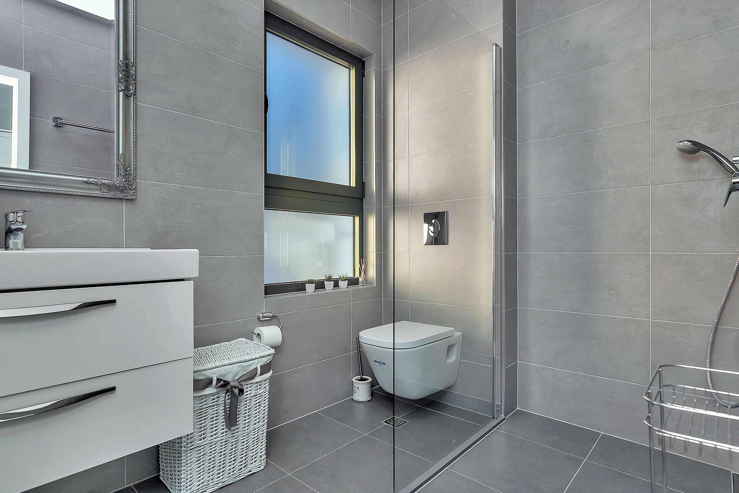 A modern bathroom with grey tiles, a white toilet, a glass shower enclosure, and a window with a view.