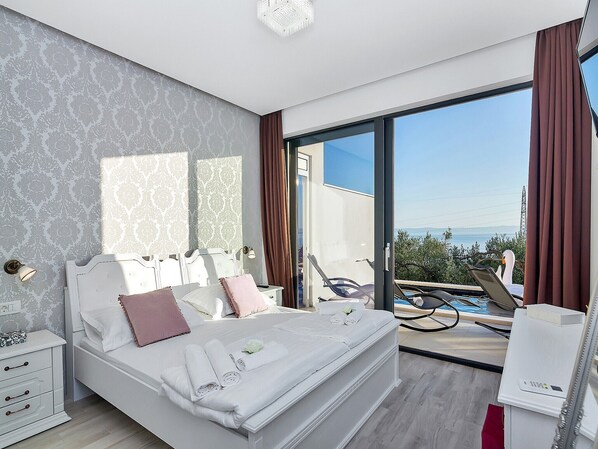 A modern bedroom with a large bed, a view of the sea, and a pool area visible through the glass door.