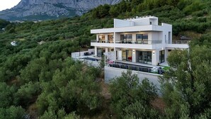 Une maison moderne à deux étages, dotée d’un balcon, entourée d’une végétation luxuriante et avec des montagnes en toile de fond.