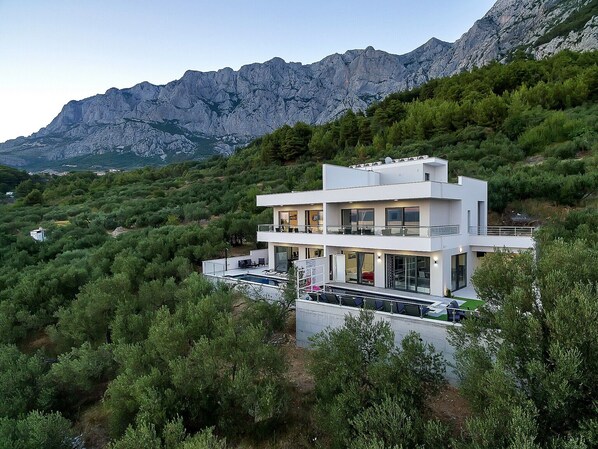 A modern two-story house with a balcony, surrounded by dense greenery and mountains in the background.