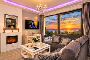 A modern living room with a fireplace, a television, a sofa, and a large window with a sunset view. - Modern holiday house Anja with heated pool and sea view, 3 bedrooms 6+2 pers. (Makarska)