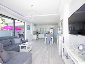 A modern living room with a grey sofa, a coffee table, and a dining area with a white table and chairs. A kitchen with white cabinets and a chandelier is visible in the background.
