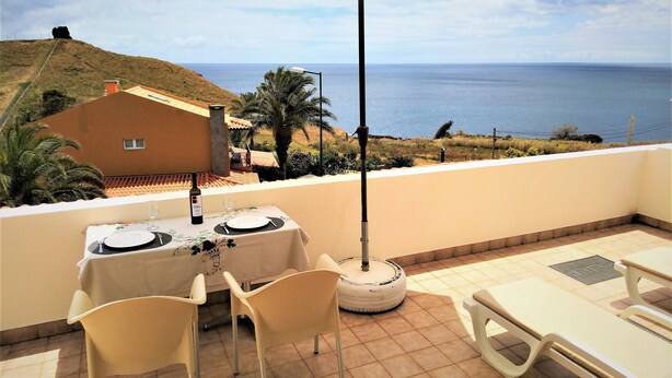 A balcony with a table set for two, a chair, and a sun umbrella. The view includes the sea and a hill in the background.