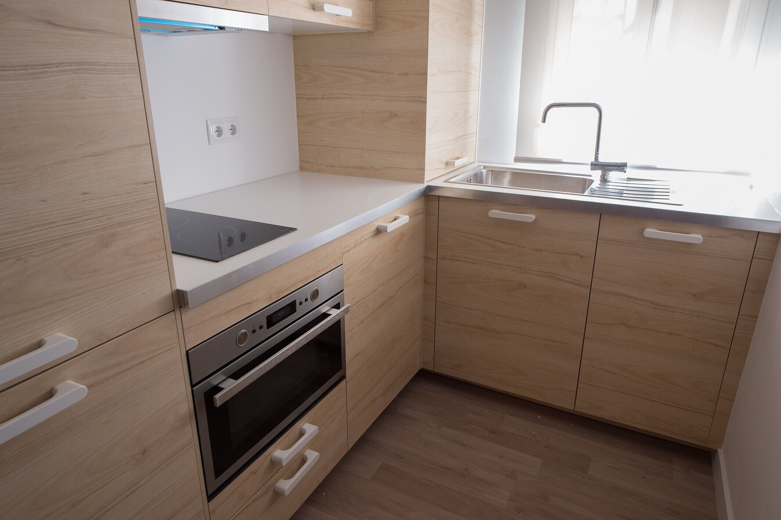 A compact kitchen with wooden cabinets, a built-in oven, a dishwasher, and a window with a sink.