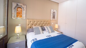 Un dormitorio con cabecera capitonada, dos mesitas de noche con lámparas, cama con ropa de cama estampada y dos cuadros en la pared.