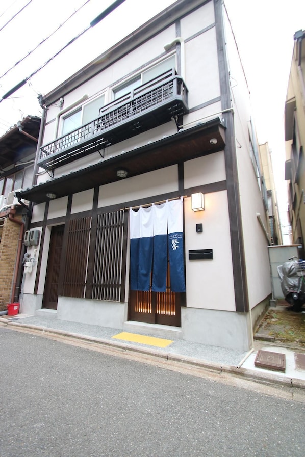 A two-story building with a balcony, a sliding door with a blue curtain, and a small step leading to the entrance.