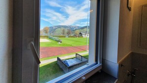 A window with a view of a green field and a red track, a small table with a book, and a door with a towel rack. - Panorama App 2C am See, für 2-4,free WIFI (Zell am See)