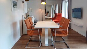 A modern dining area with a white table and orange chairs, a framed picture on the wall, and a TV mounted on the wall. - Panorama App 2C am See, für 2-4,free WIFI (Zell am See)