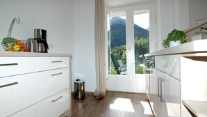 Una cocina moderna con armarios blancos, una cafetera de acero inoxidable y un cubo de basura. Se ve una vista de sillas verdes y una montaña a través de la puerta.