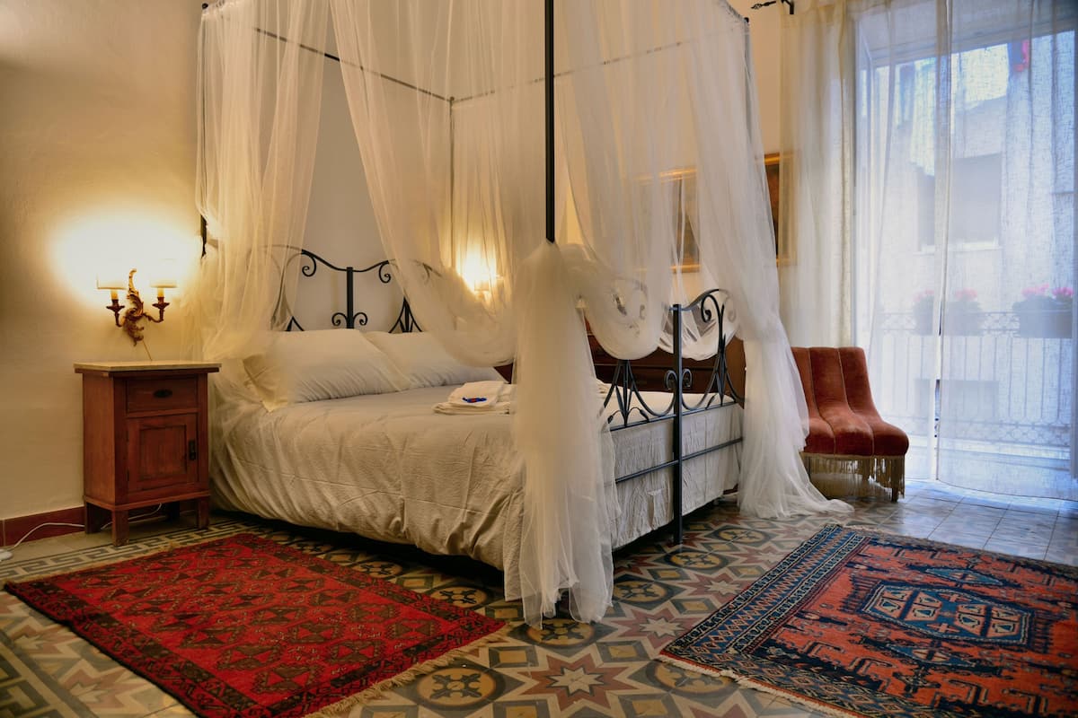 A four-poster canopy bed with a nightstand and lamp, a red and blue patterned rug, and a red armchair.