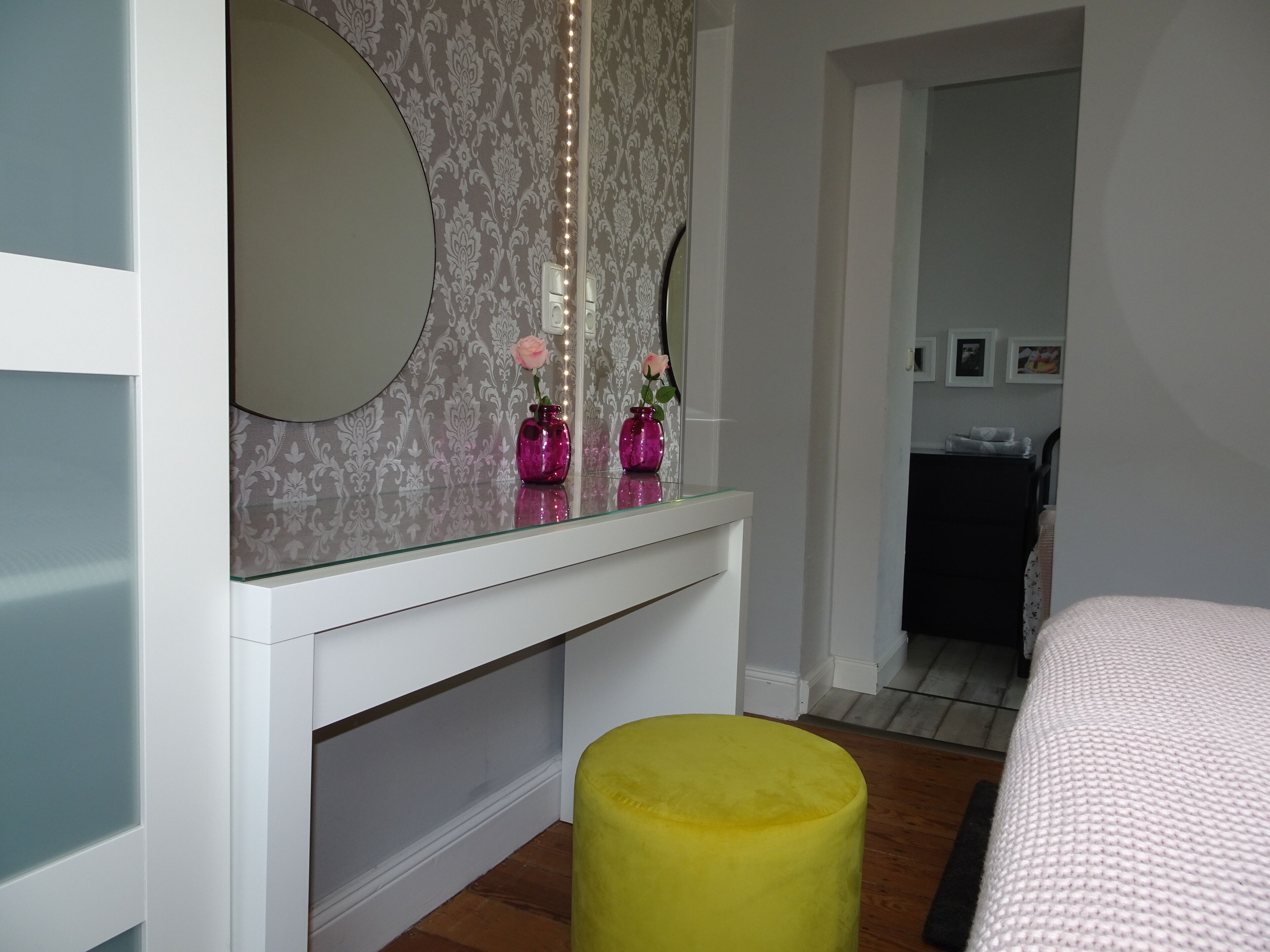 A bedroom with a bed, a vanity with a mirror, and a yellow ottoman.