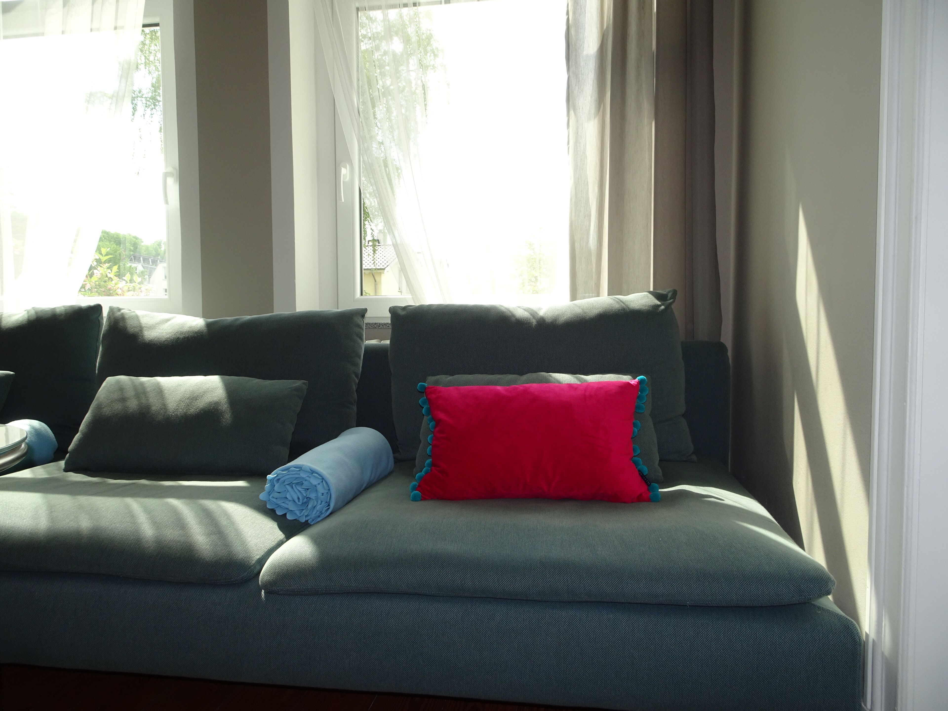A sofa with multiple cushions, a window with curtains, and a view of greenery outside.
