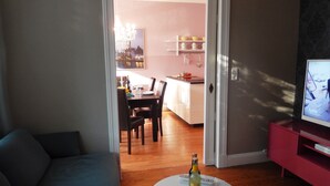 A living room with a sofa, a round coffee table with a bottle, a TV on a stand, and a dining area in the background.