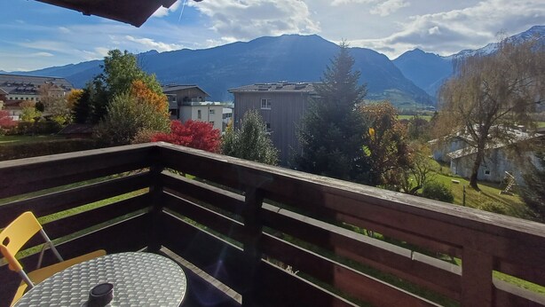 En balkon med et bord og stole, med udsigt over et bjerglandskab med bygninger og træer.