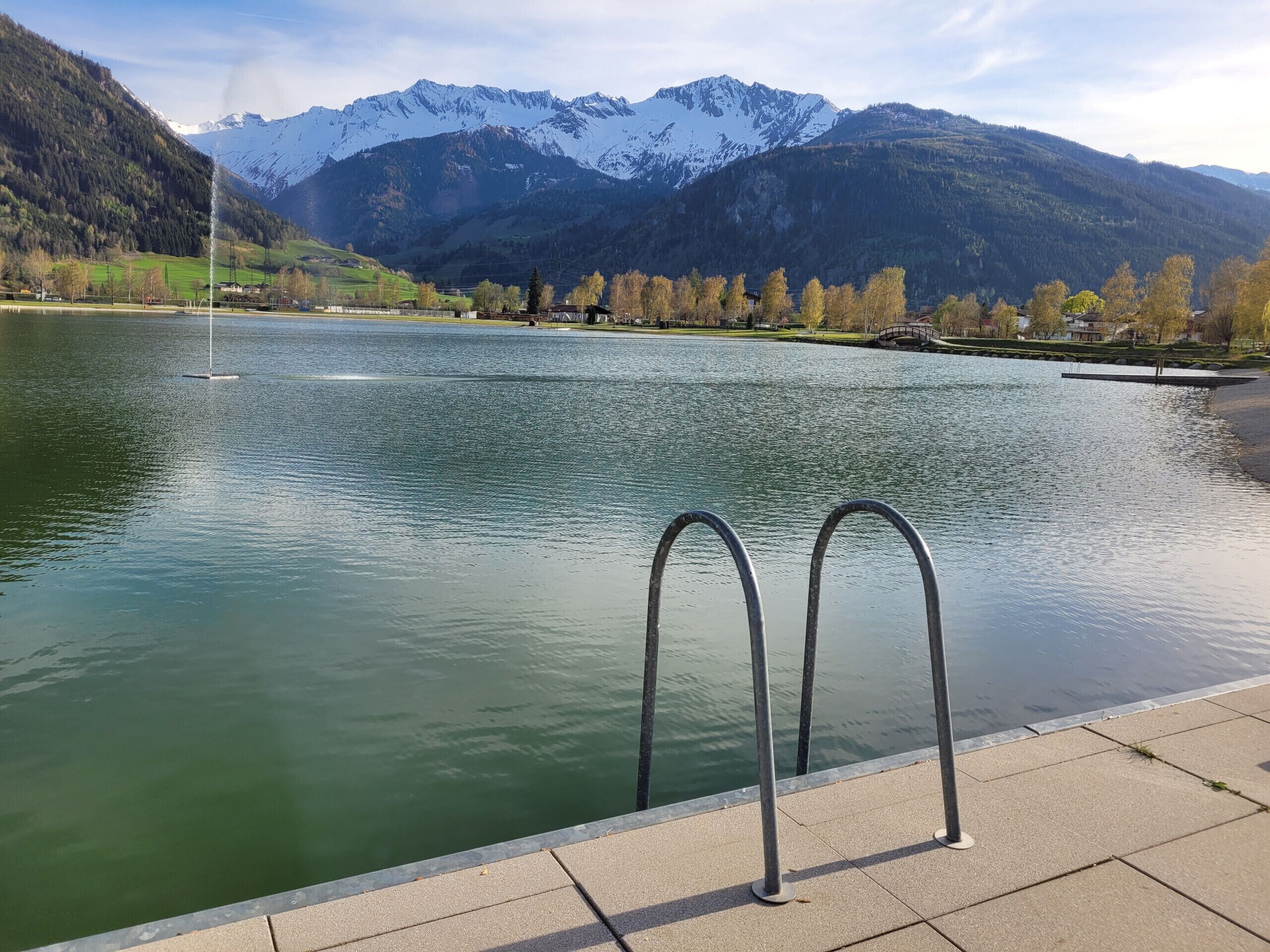 A lake with mountains in the background, a fountain, and a metal railing along the edge.