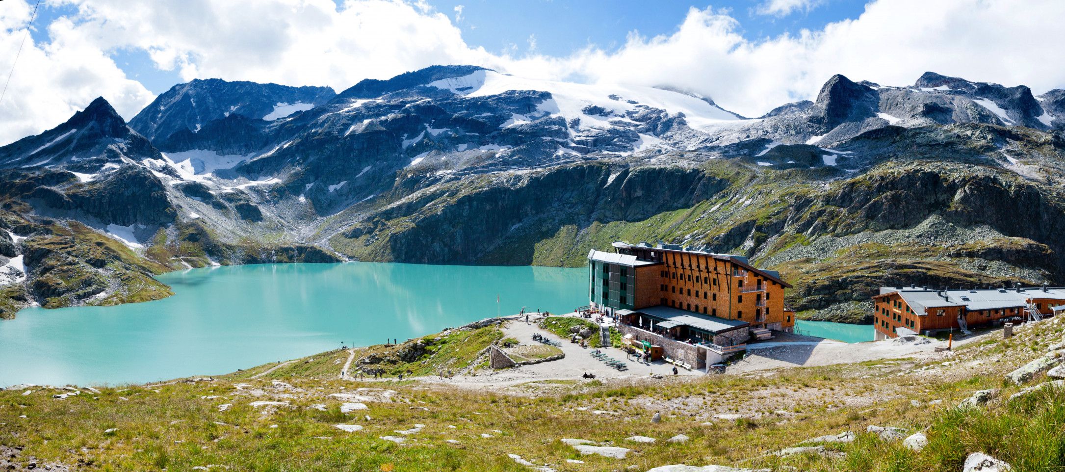Un chalet de montagne doté d’une grande façade en verre, avec un lac et des montagnes enneigées en arrière-plan.
