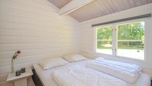 A bedroom with white wooden walls, a bed with white bedding, a window with a view of greenery, and a bedside table with a vase and two candle holders. - 3 bedroom gorgeous home in Gedser (Gedser)