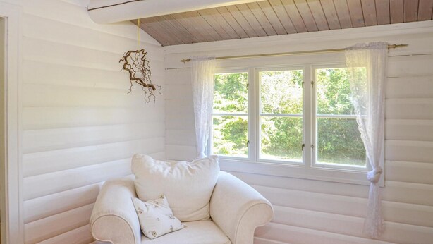 A white armchair with a cushion and a small table in a room with wooden walls and a large window with curtains. - 3 bedroom gorgeous home in Gedser (Gedser)