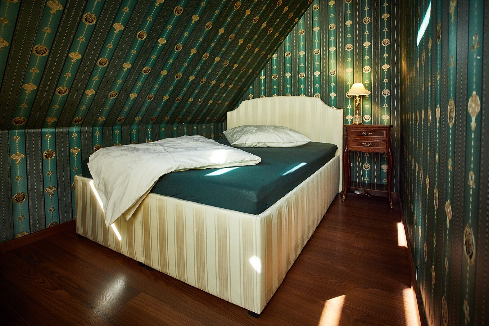 Ein Schlafzimmer mit schrägen Wänden, einem Bett mit gestreifter Kopfteil, einem Nachttisch mit Lampe und Wandtapete mit Muster.