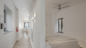 Une chambre d’hôtel moderne au design minimaliste, dotée d’un plancher en bois, d’un lit avec une tête de lit intégrée, d’un ventilateur de plafond et d’une fenêtre avec des rideaux.