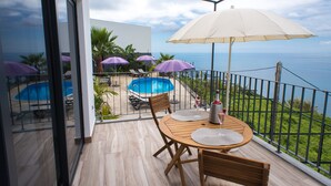 Un balcon avec une table en bois, des chaises et un grand parasol. Il y a une piscine et une vue sur l’océan.