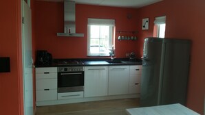 A kitchen with white cabinets, a stainless steel refrigerator, a stove, and a window with a view of the outside. - Modern house, 6 persons, 3 bedrooms, 2 bathrooms, smoke free, TOP location (De Cocksdorp)