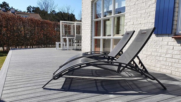 A deck with two lounge chairs and a table, a white brick building with large windows, and a hedge in the background. - Modern house, 6 persons, 3 bedrooms, 2 bathrooms, smoke free, TOP location (De Cocksdorp)