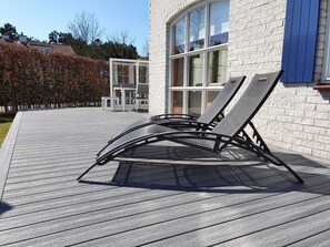 A deck with two lounge chairs and a table, a white brick building with large windows, and a hedge in the background. - Modern house, 6 persons, 3 bedrooms, 2 bathrooms, smoke free, TOP location (De Cocksdorp)