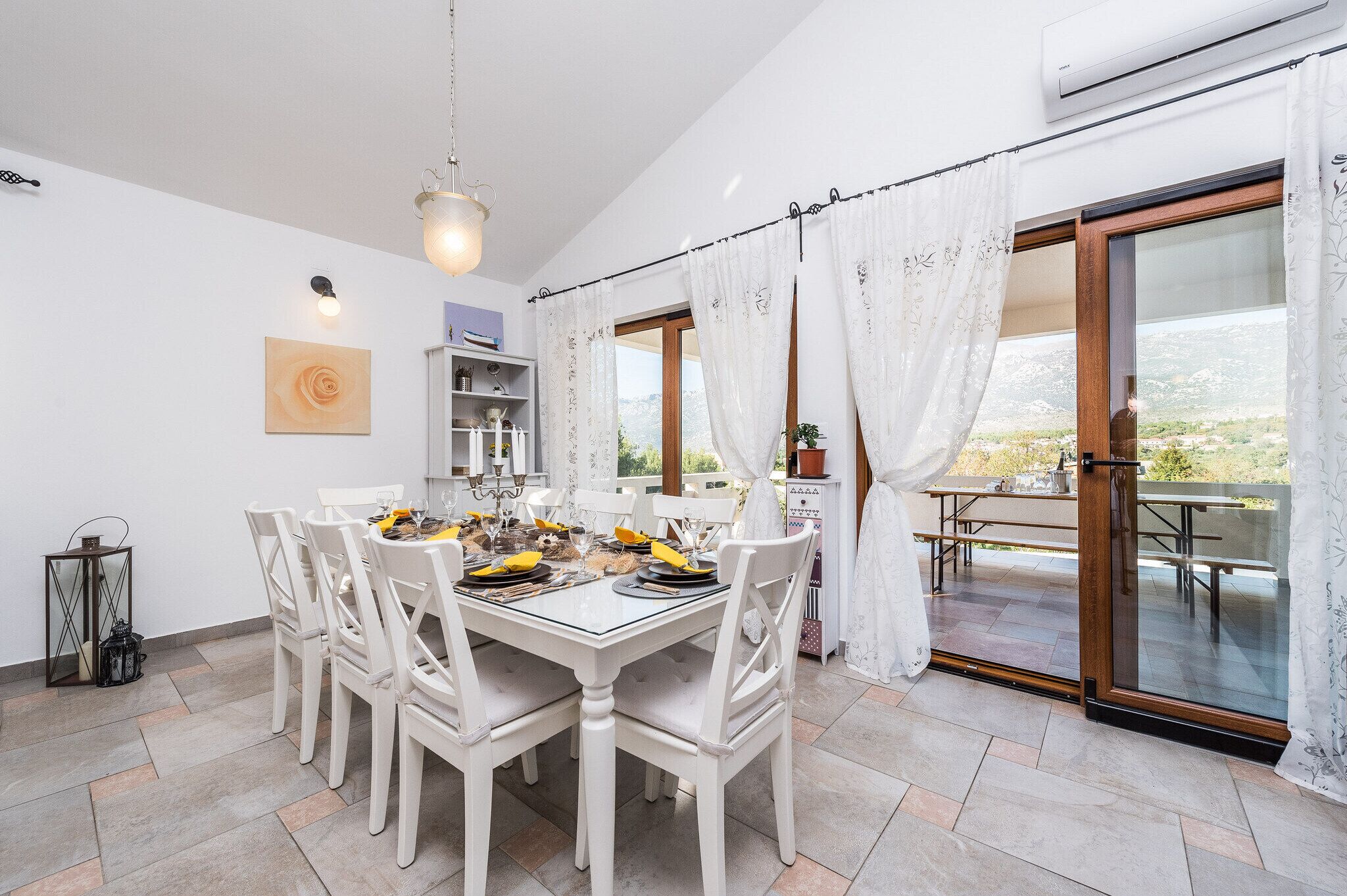 A dining area with a white table set for six, a chandelier, and a view of a balcony with outdoor seating.