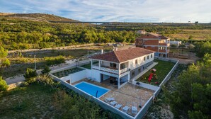 Una casa moderna de dos pisos con piscina, rodeada de vegetación y colinas.