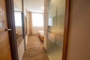 A hotel room with a wooden floor, a bed with white bedding, a large mirror, and a glass partition wall.