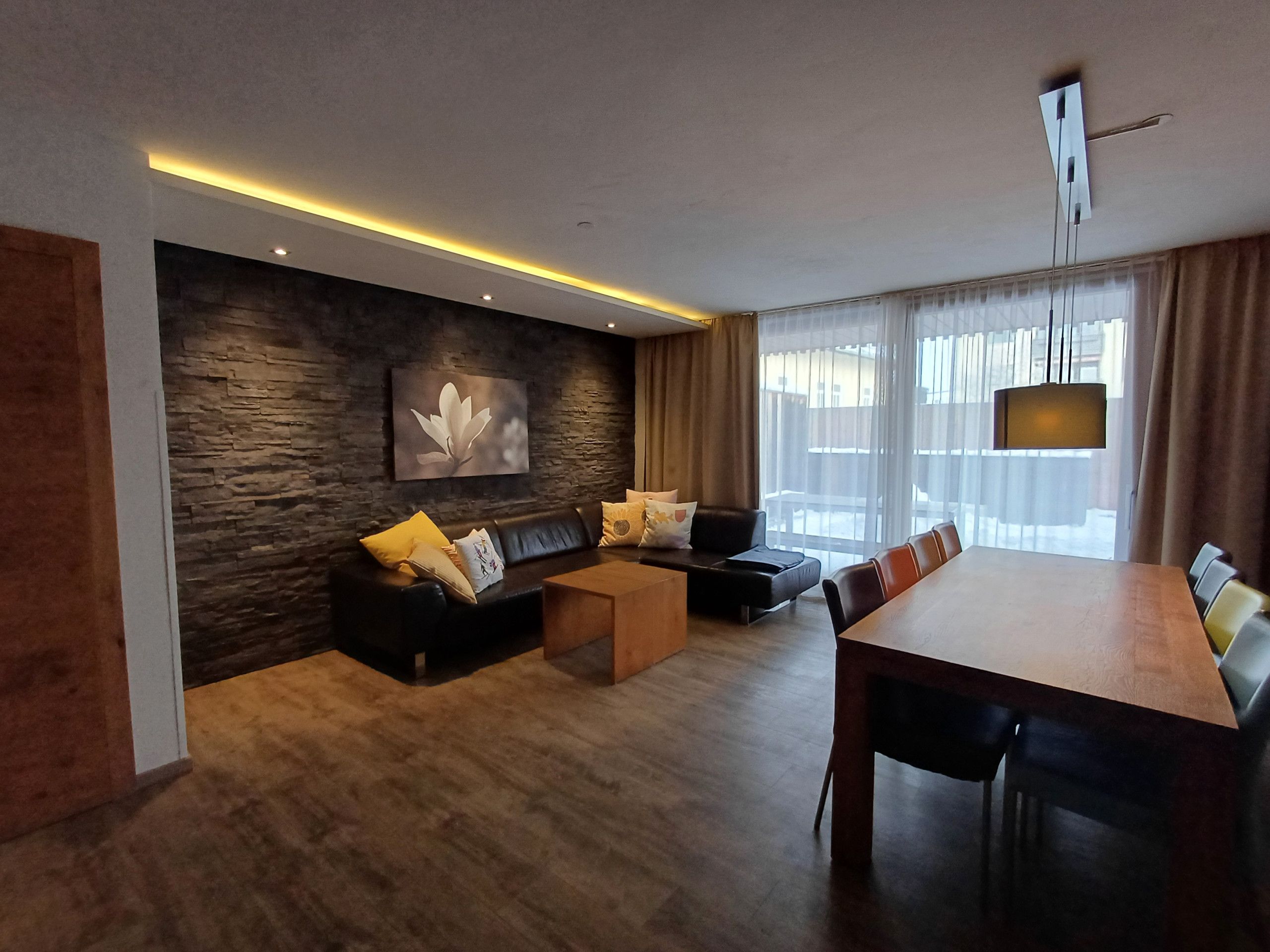 A modern living room with a stone accent wall, a black leather sofa, a wooden coffee table, and a dining area with a long table and chairs.