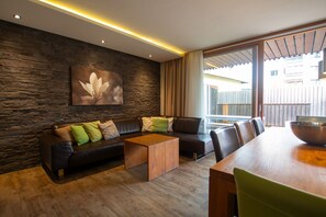 A modern living room with a stone wall, a black leather sofa with green cushions, a wooden coffee table, and a dining table with chairs.