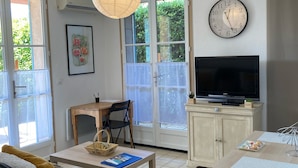 A living room with a sofa, a dining table with chairs, a TV, a clock, and a window with a view of greenery.