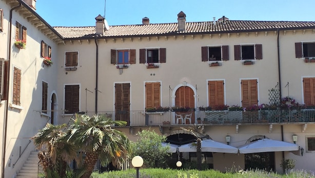 Un bâtiment doté de nombreuses fenêtres, dont certaines sont équipées de volets, un balcon avec des chaises et un petit jardin agrémenté d’un palmier et d’un lampadaire.