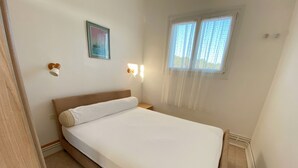 Una cama individual con cabecera de madera, una mesita de noche, una lámpara de pared, una ventana con cortinas traslúcidas y un cuadro enmarcado en la pared.