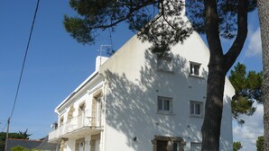 Un edificio blanco de dos pisos con un balcón en el segundo nivel, fachada de piedra y un árbol grande al frente.