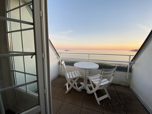 Varanda com mesa e cadeiras brancas, com vista para um corpo de água ao pôr do sol.