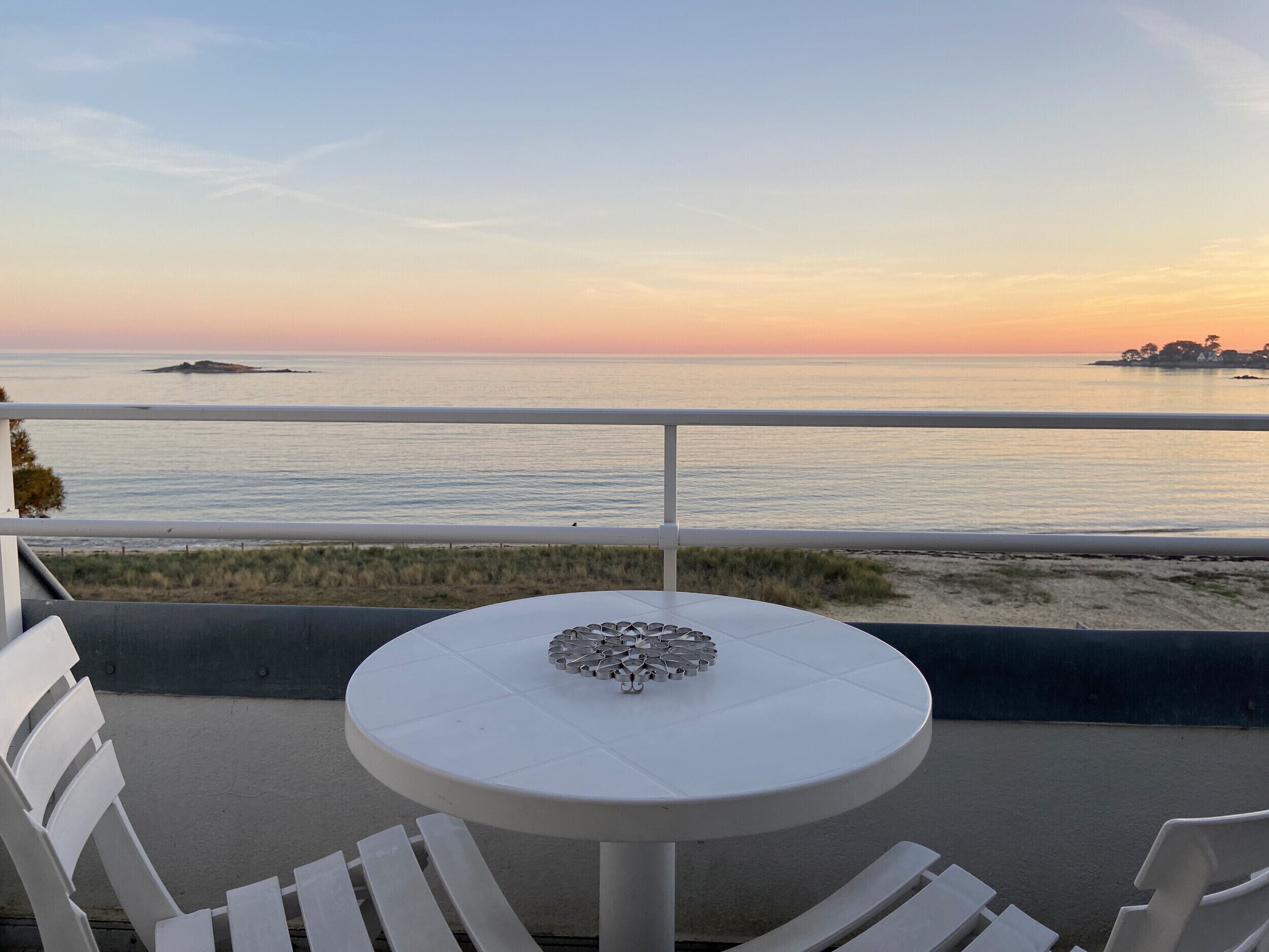 A round table with a decorative centerpiece, two white chairs, and a view of the ocean at sunset.