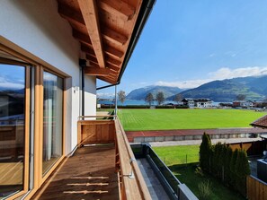 Un balcón con piso de madera, puerta de cristal y vista a un campo verde y montañas al fondo.