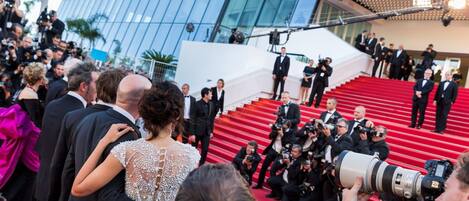 A red carpet event with people in formal attire, a modern building with glass windows, and photographers capturing the event.