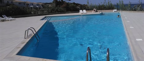 A rectangular swimming pool with clear blue water, surrounded by a tiled deck. There are lounge chairs and a small table on the deck.