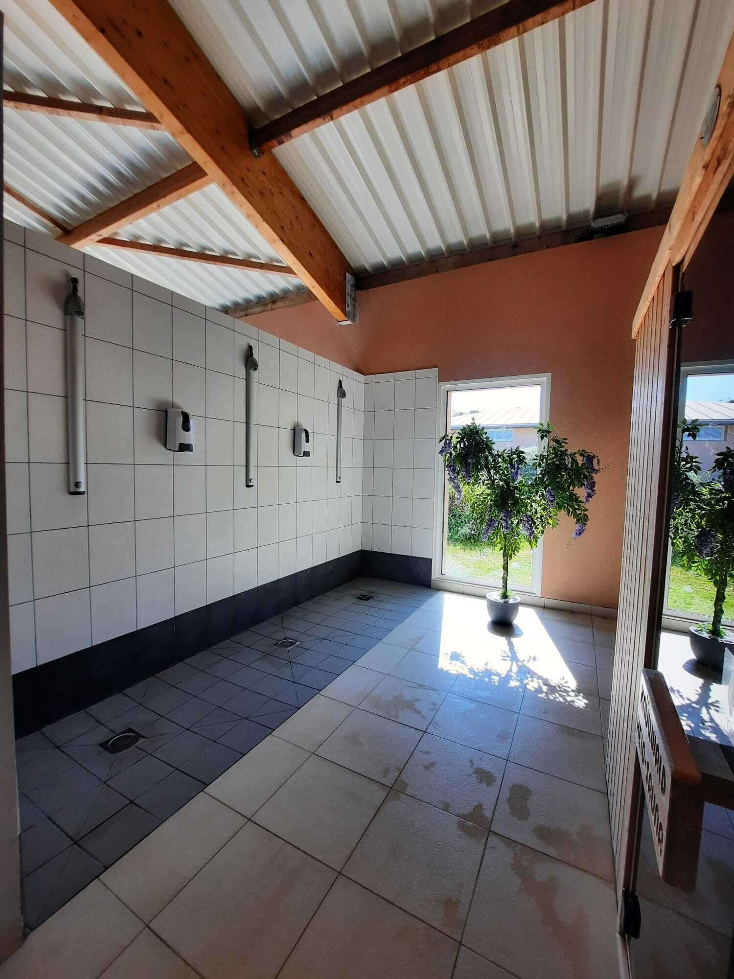 Une cuisine spacieuse avec des murs recouverts de carreaux blancs, un sol carrelé et un plafond en bois. On y trouve trois robinets fixés au mur et une plante en pot près de la fenêtre.