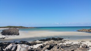 A beach with clear blue water, rocky shore, and a distant green hill.
