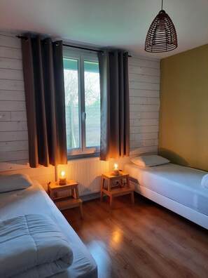 A cozy room with two beds, a wooden floor, a window with curtains, a radiator, and a hanging light fixture.