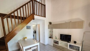 A modern living room with a wooden staircase, a dining table with chairs, a kitchen area, and a TV on a cabinet. - Garden with barbecue, swimming pool (LA SEYNE SUR MER)