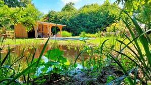 A wooden structure with a covered seating area, a pond, and lush greenery. - Charming bergerie *** gîte & spa (Pouydesseaux)